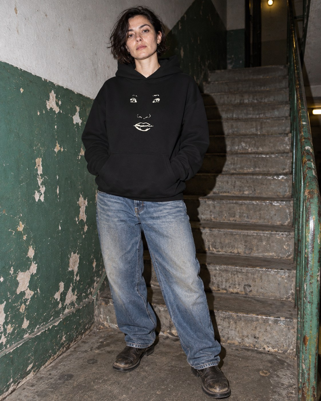 Black Face hoodie with cream face drawing, styled with jeans in a stairwell