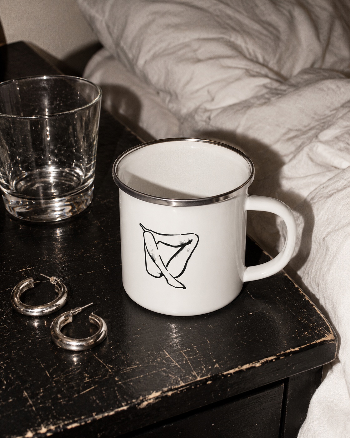 White Folded enamel mug with black drawing and silver rim on a worn bedside table