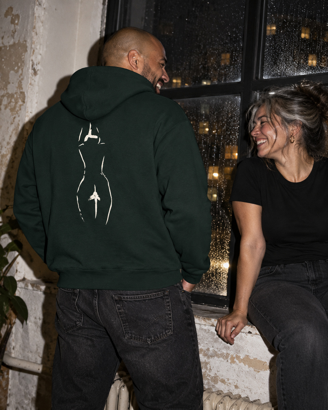 Pine Green Turn Away hoodie with cream back drawing, worn by a man laughing with a friend beside a rainy window