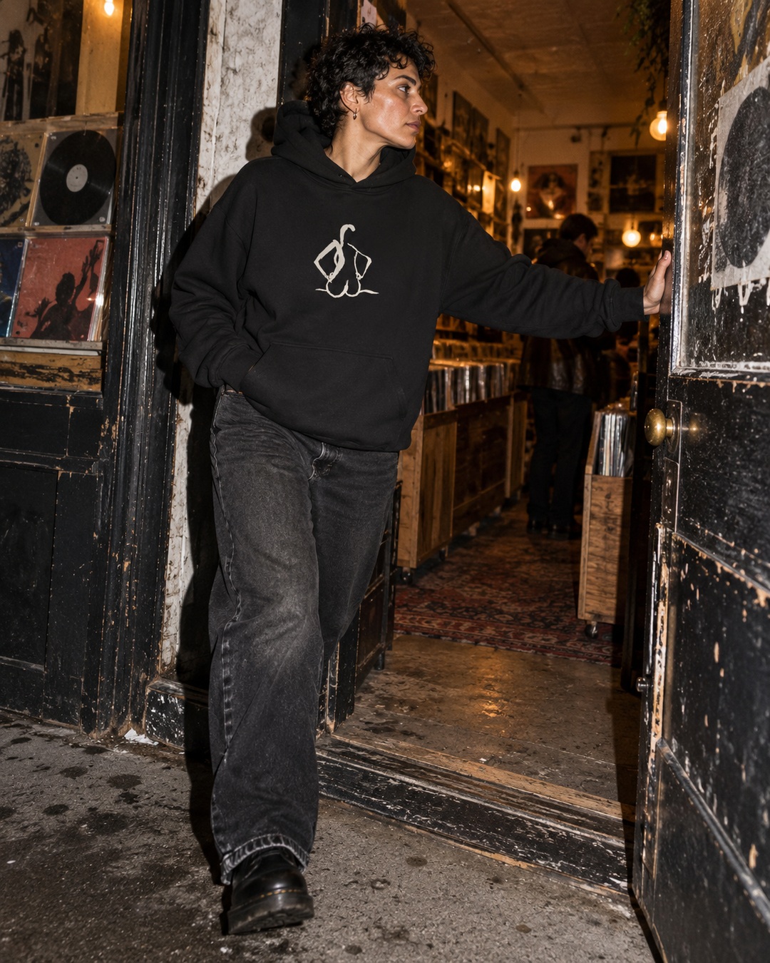 Black Hands on Hips hoodie with cream chest drawing, worn while stepping through a worn record-shop doorway