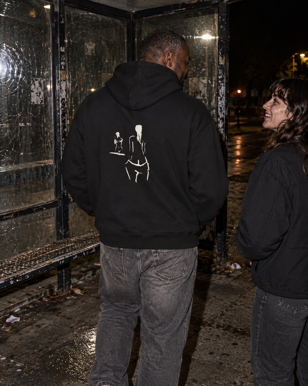 Black On Reflection hoodie with cream back drawing worn by a man talking with a friend at a worn bus shelter at night