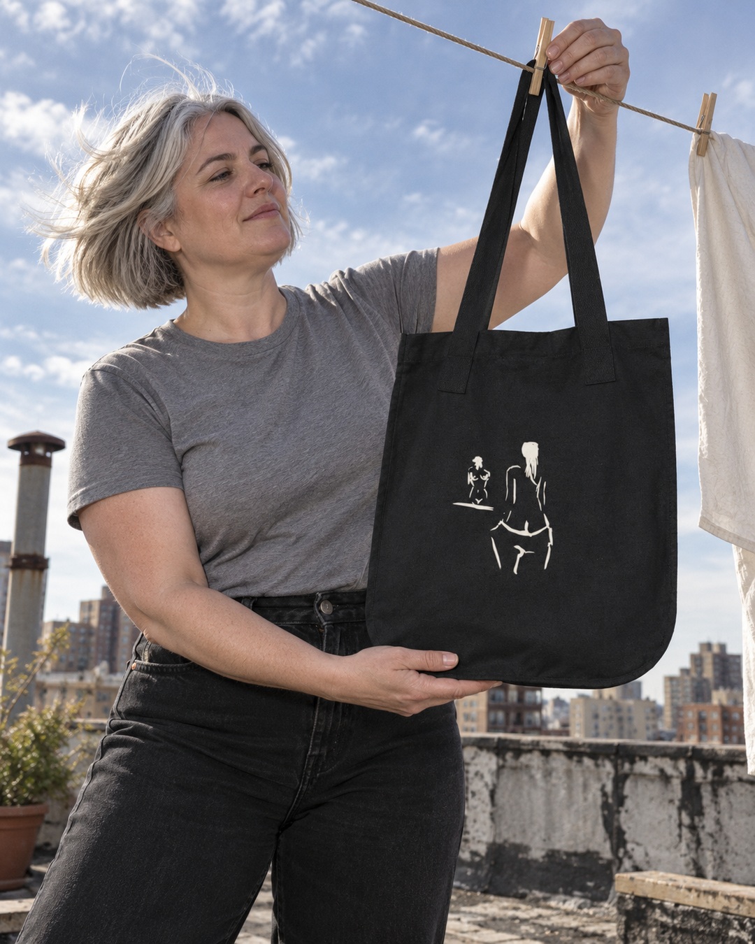 Black On Reflection tote with cream ink drawing held beneath a rooftop clothesline by a silver-haired woman