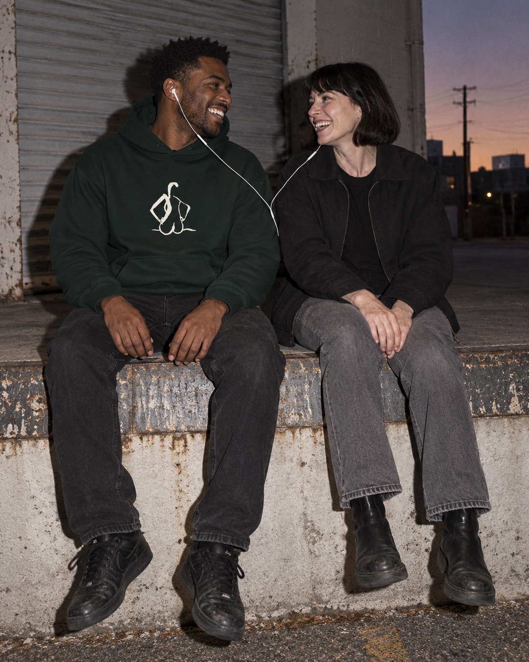 Pine Green Hands on Hips hoodie with cream chest drawing worn by a man sharing earbuds with a friend on a worn loading platform