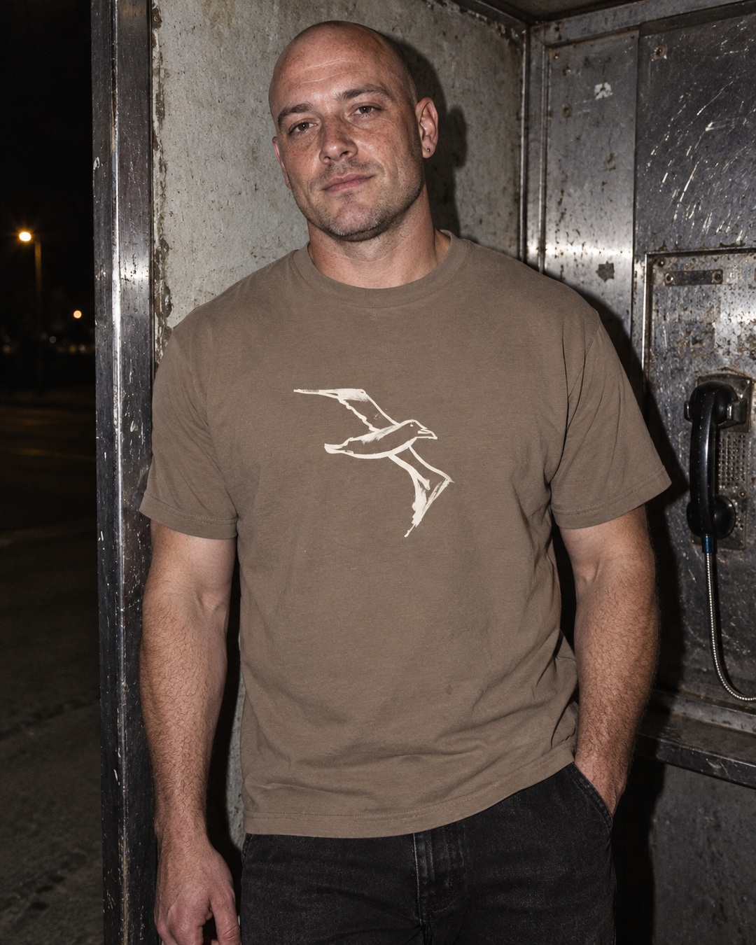 Faded Brown Albatross heavy tee with cream bird drawing worn by a man in a scuffed telephone alcove
