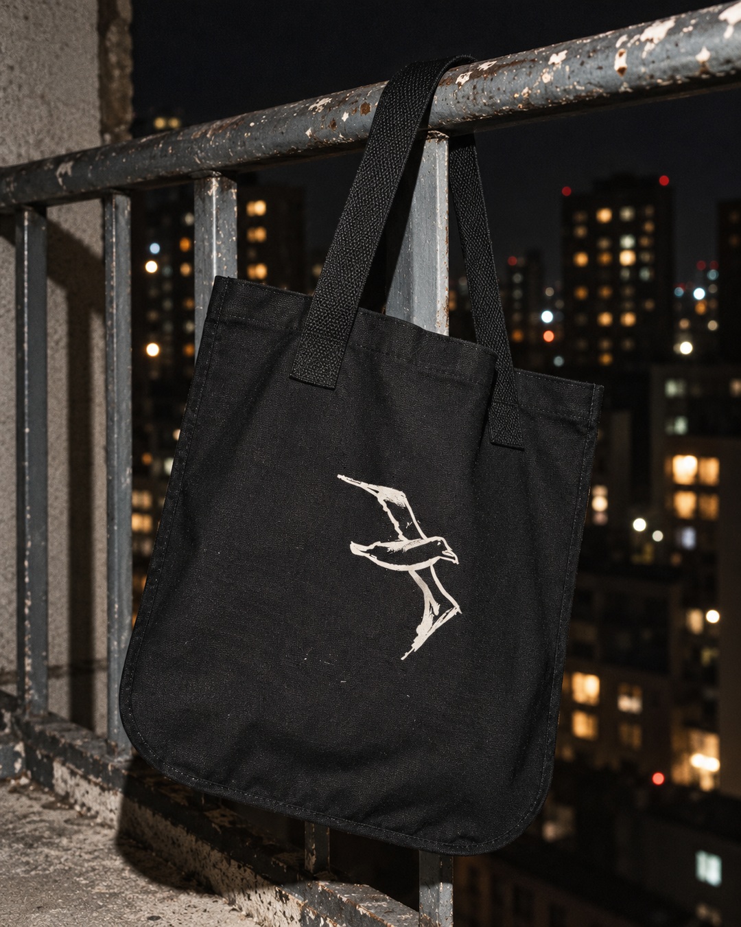 Black Albatross canvas tote with cream bird drawing hanging from a weathered balcony railing at night