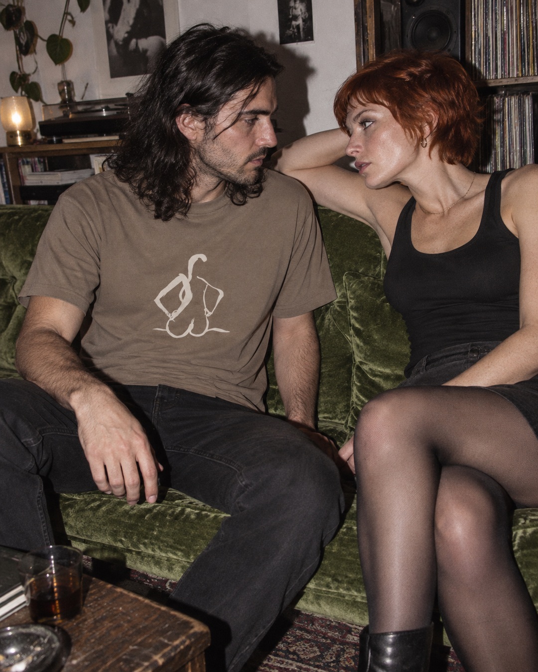 Faded Brown Hands on Hips tee with cream ink drawing, worn beside a woman on a green velvet sofa
