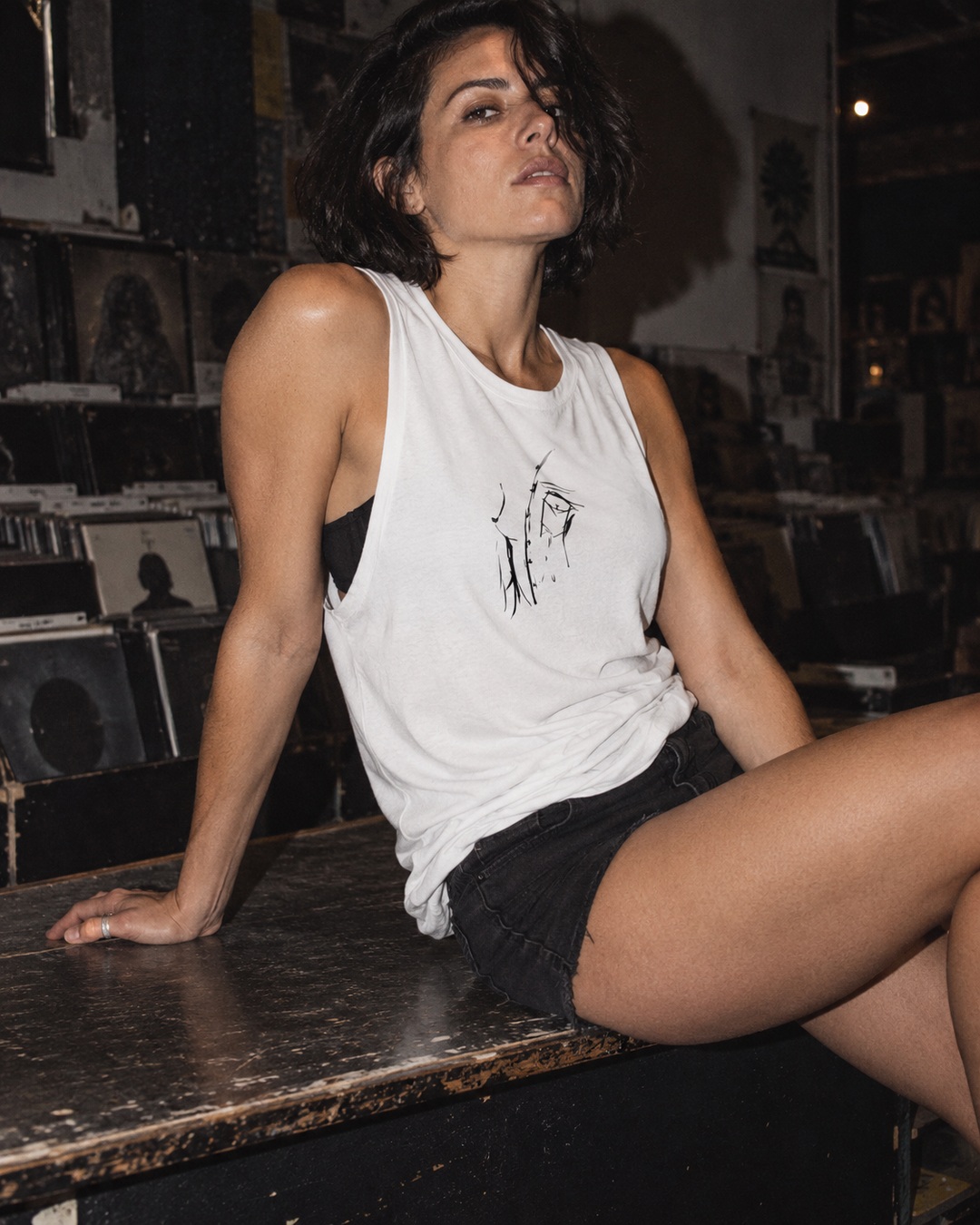 White Peaking Out raw tank with black ink artwork, worn with black denim shorts on a scuffed record-store counter