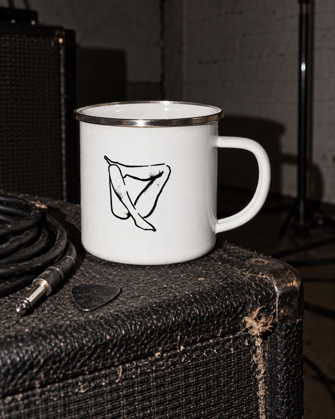 White Folded enamel mug with black ink drawing and silver rim on a worn amplifier beside an instrument cable