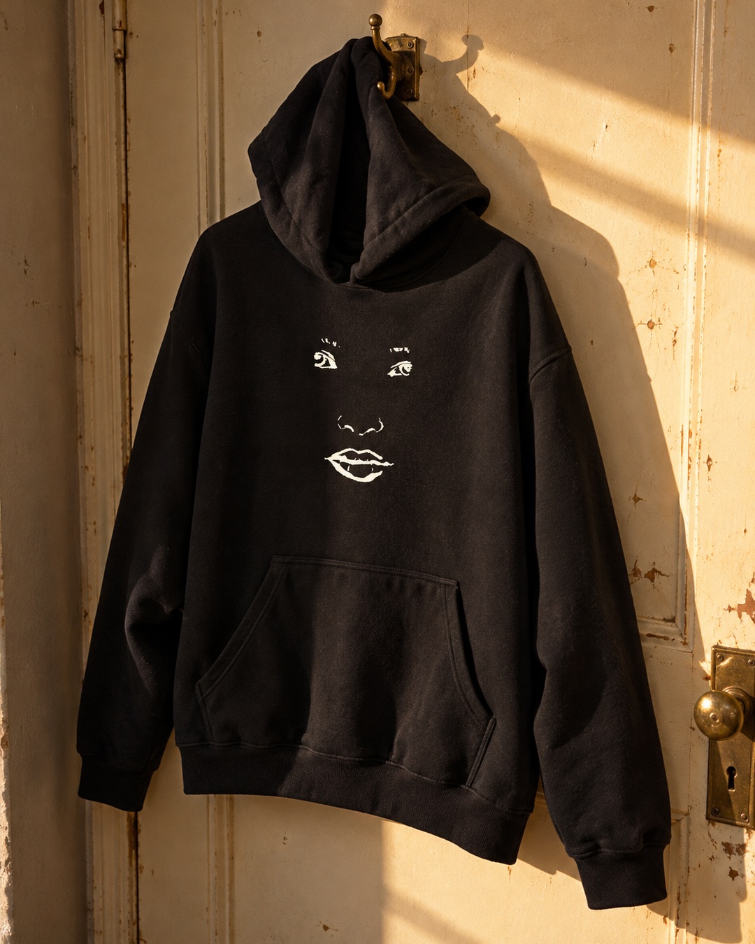Black Face hoodie with cream face drawing hanging from a brass hook on a sunlit worn door