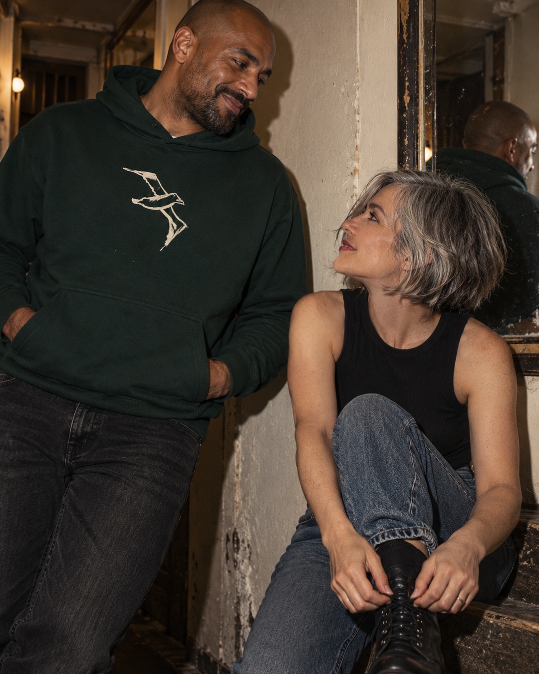 Pine Green Albatross hoodie with cream bird drawing worn beside a friend tying a boot in a worn hallway