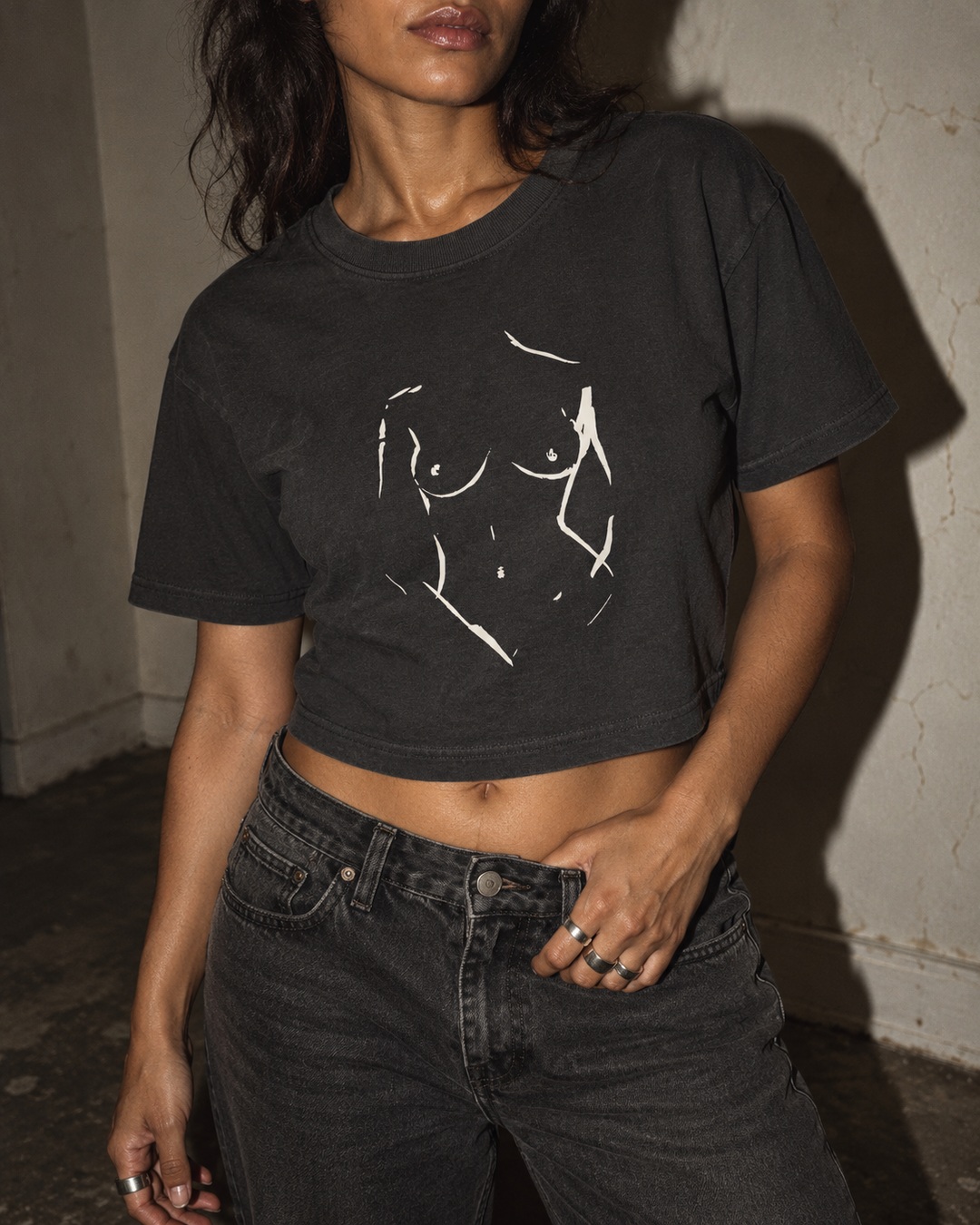 Faded Black Bare Form cropped tee with cream ink drawing, worn with charcoal jeans against a cracked cream wall