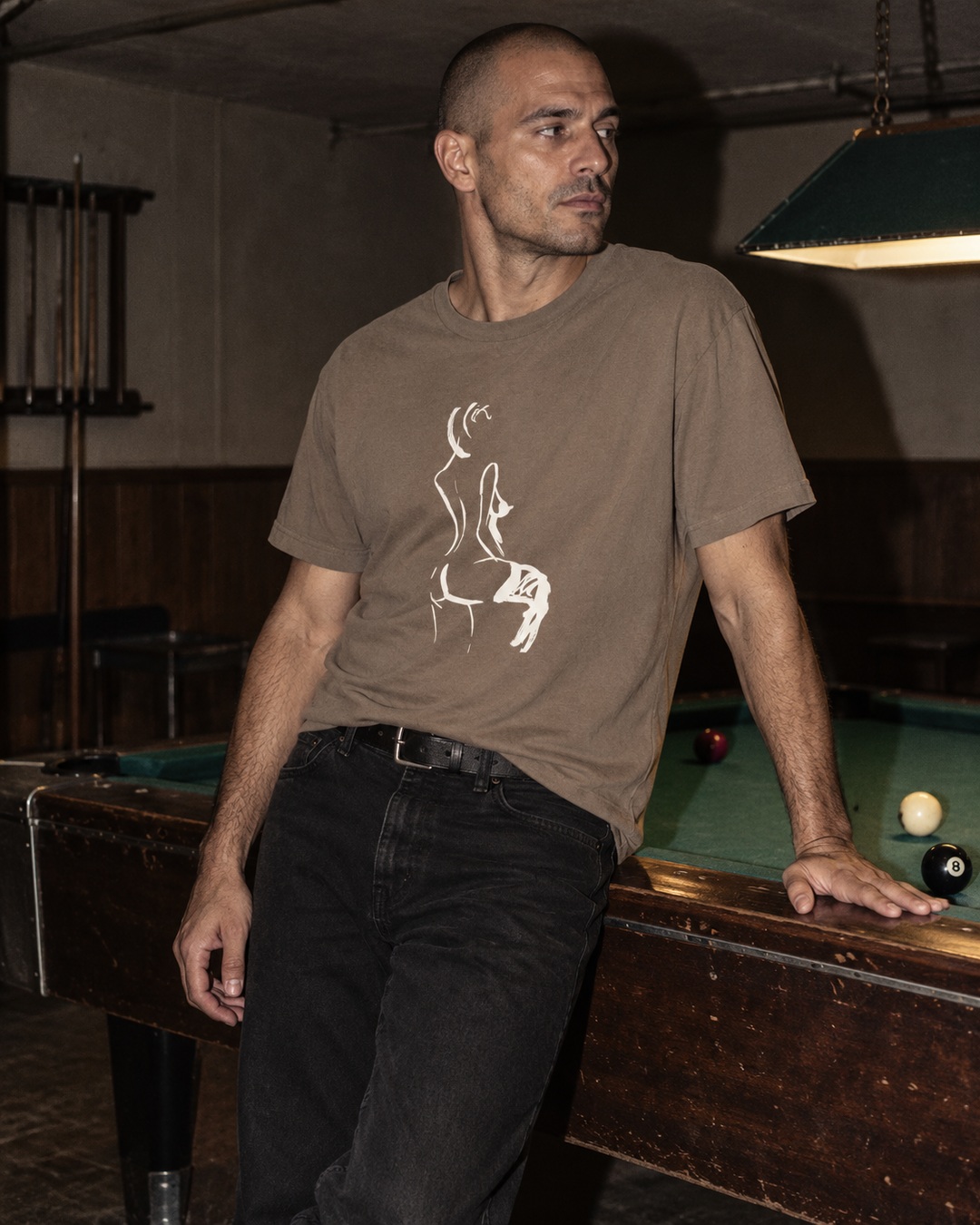 Faded Brown Seated heavy tee with cream ink drawing worn by a man leaning against a worn pool table